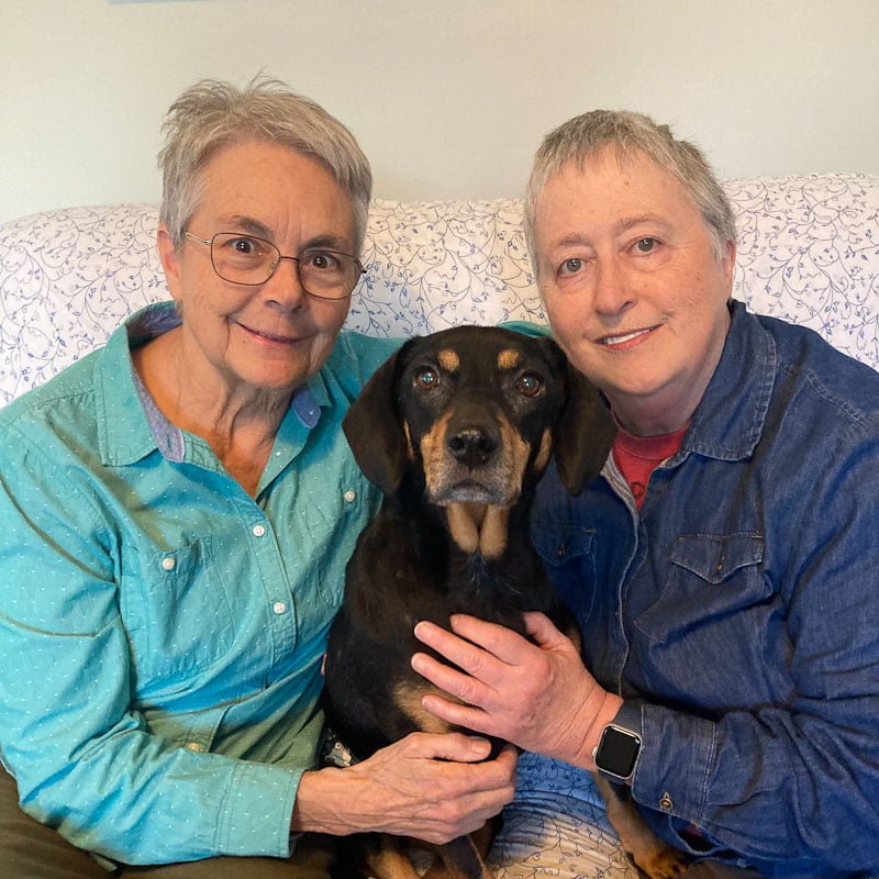 Two women one with glasses and a light blue shirt and the other with a dark blue shirt are sitting on a couch with a black dog sitting between them.