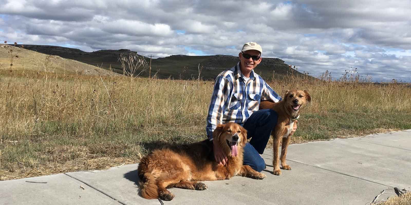 A man and two dogs, one a shepherd mix and the other a terrier mix on a sidewalk on a prairie with view of hills in the background.