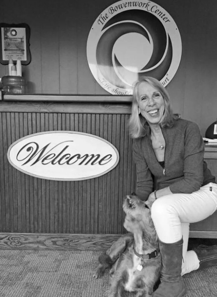 A smiling woman with a grey sweater and white pants is kneeling by a welcome sign with a scruffy dog who has his head turned up to her.