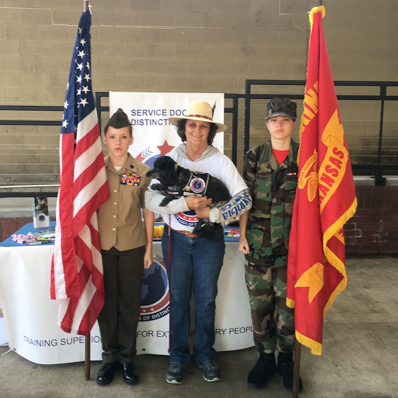 A woman with a white cowboy had in long sleeve shirt and jeans holds her black/white dog Bailey and they are flanked by young military members holding the US flag and State of Arkansas flag.